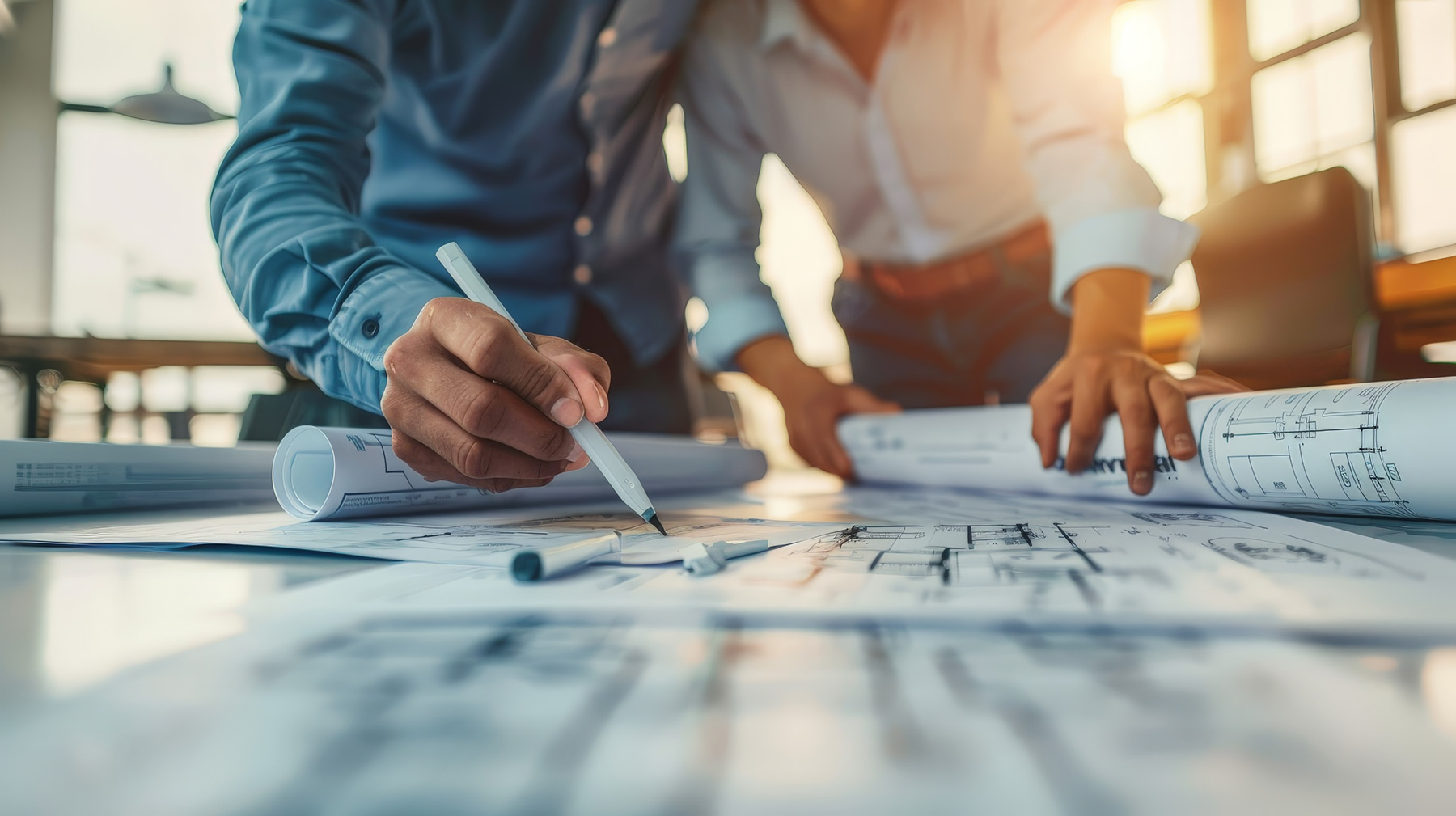 Zwei Personen arbeiten zusammen an Bauplänen, einer hält einen Stift und markiert Details auf den Plänen, während der andere ein gerolltes Papier festhält. Das Bild ist von warmem Licht durchflutet.