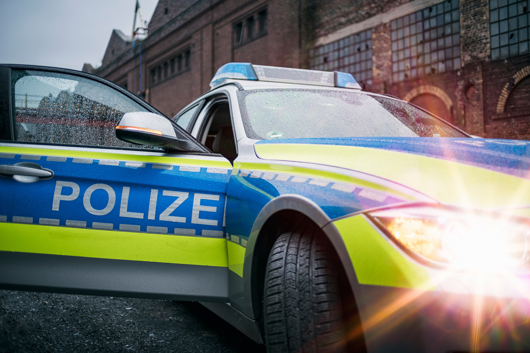 Ein Polizei-Auto mit blau-grüner Beschriftung steht vor einer alten Fabrikgebäude. Die Fahrertür ist geöffnet, und das Blaulicht des Fahrzeugs leuchtet. Es regnet leicht, und Tropfen sind auf dem Auto sichtbar.